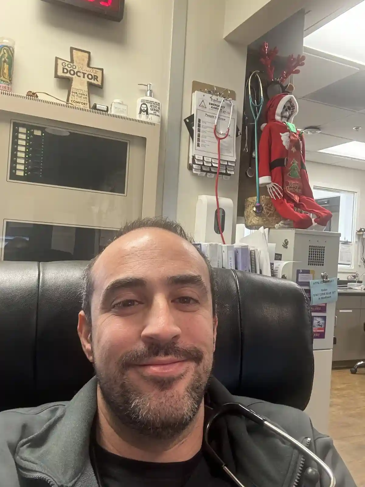 A man with a stethoscope around his neck smiles at the camera from a doctor's office chair. Behind him, a shelf holds a…