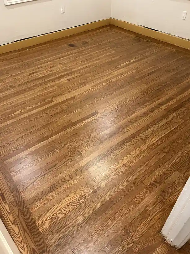 A photo shows a newly installed hardwood floor in a room with white walls and baseboards, featuring visible electrical…