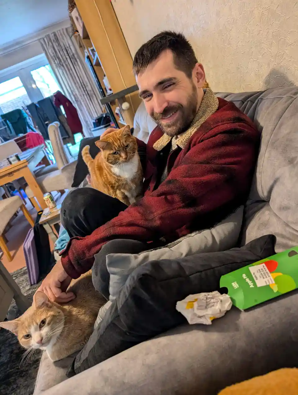A man with a beard and red jacket sits on a grey couch smiling at the camera while petting two orange and white cats. One…