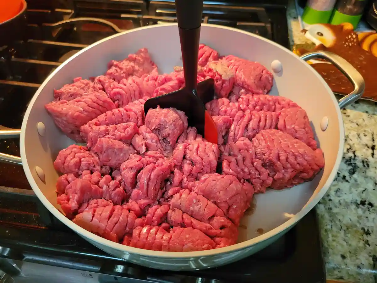 A white frying pan sits on a stovetop, filled with raw ground beef arranged in distinct, thick, uncooked patties. A black…