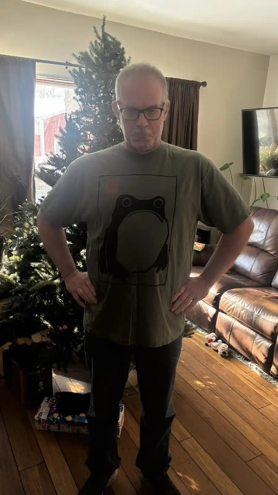 A middle-aged man with glasses stands indoors with hands on hips, wearing a green t-shirt featuring a black silhouette of a…