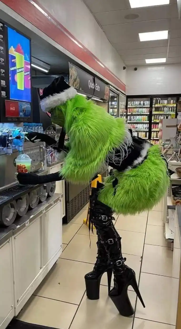 A person dressed as the Grinch in a bright green furry costume, wearing a Santa hat and towering black platform heels, is…