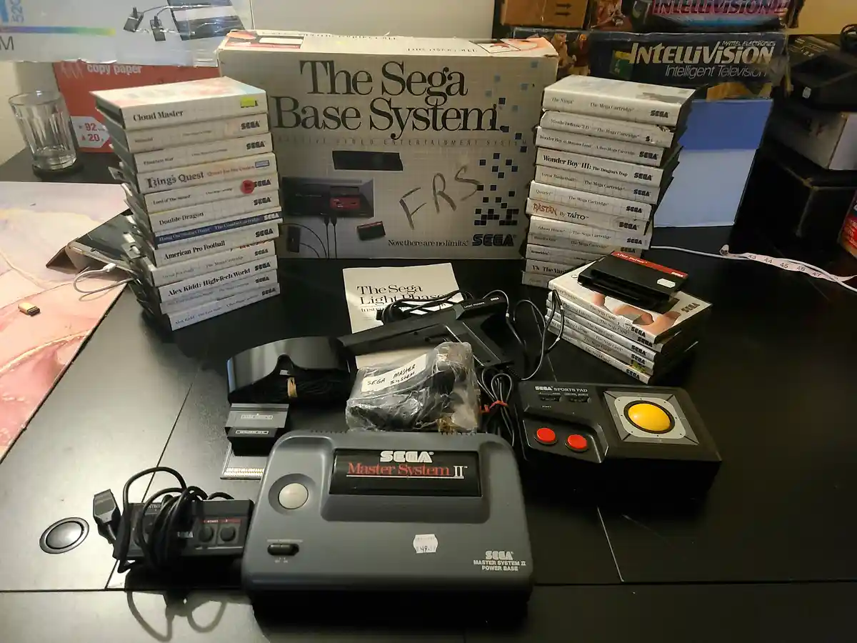 A Sega Master System II console sits on a table surrounded by stacks of its games, controllers, and accessories. Behind it…