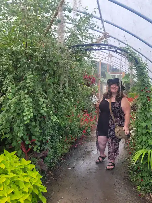 A woman stands smiling in a lush greenhouse pathway surrounded by dense green foliage and red flowers, wearing a black hat…