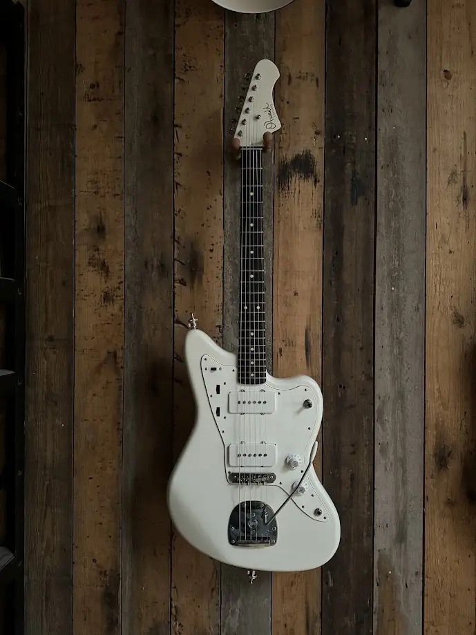 A white electric guitar, likely a Fender Jaguar, hangs vertically against a rustic wooden wall with visible grain and knots…