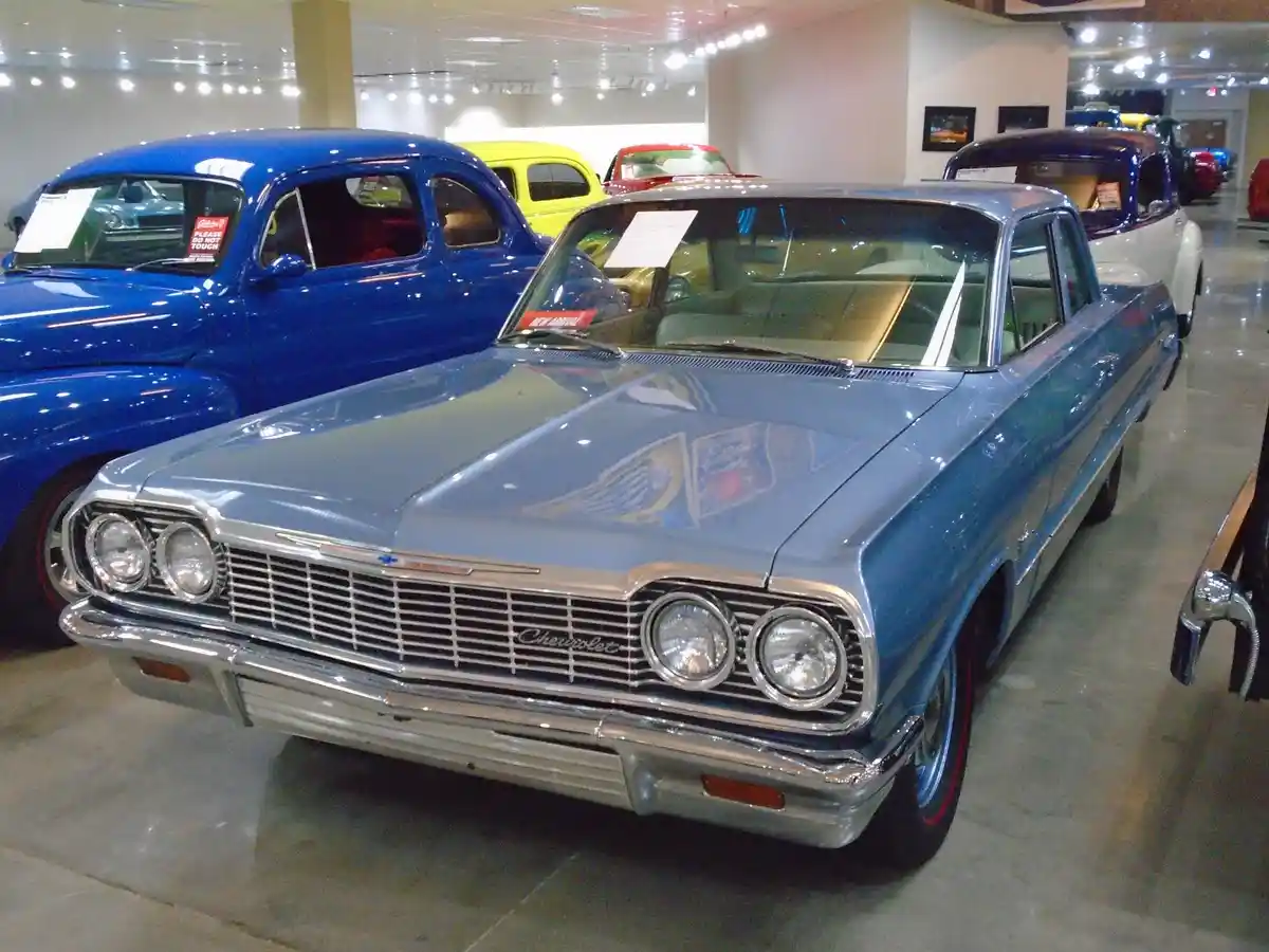 A light blue 1960s Chevrolet automobile is displayed in a museum with other vintage cars, including a blue car to its left…