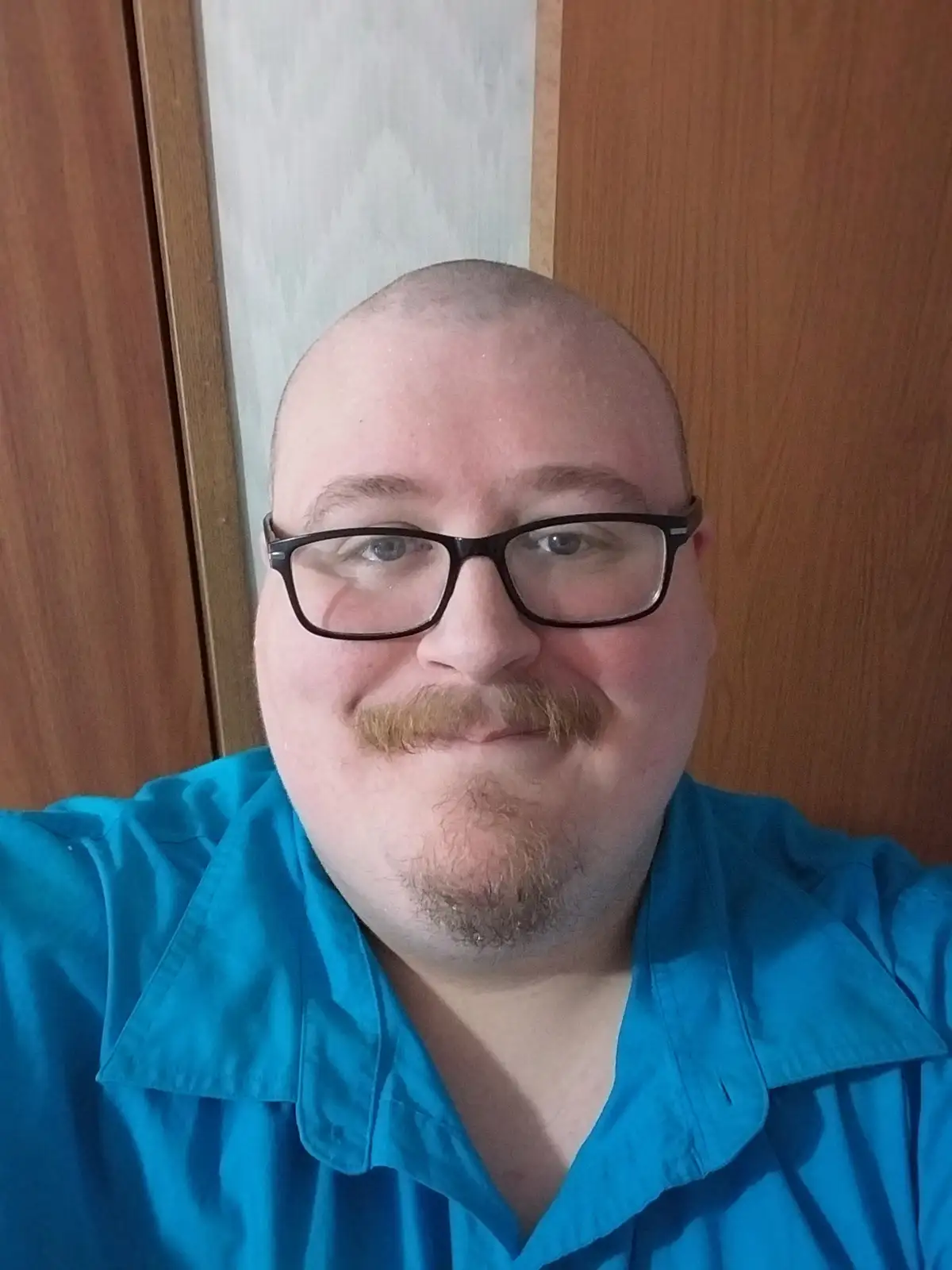 A close-up selfie of a bald man with a reddish-brown mustache and goatee, wearing black-rimmed glasses and a bright blue…