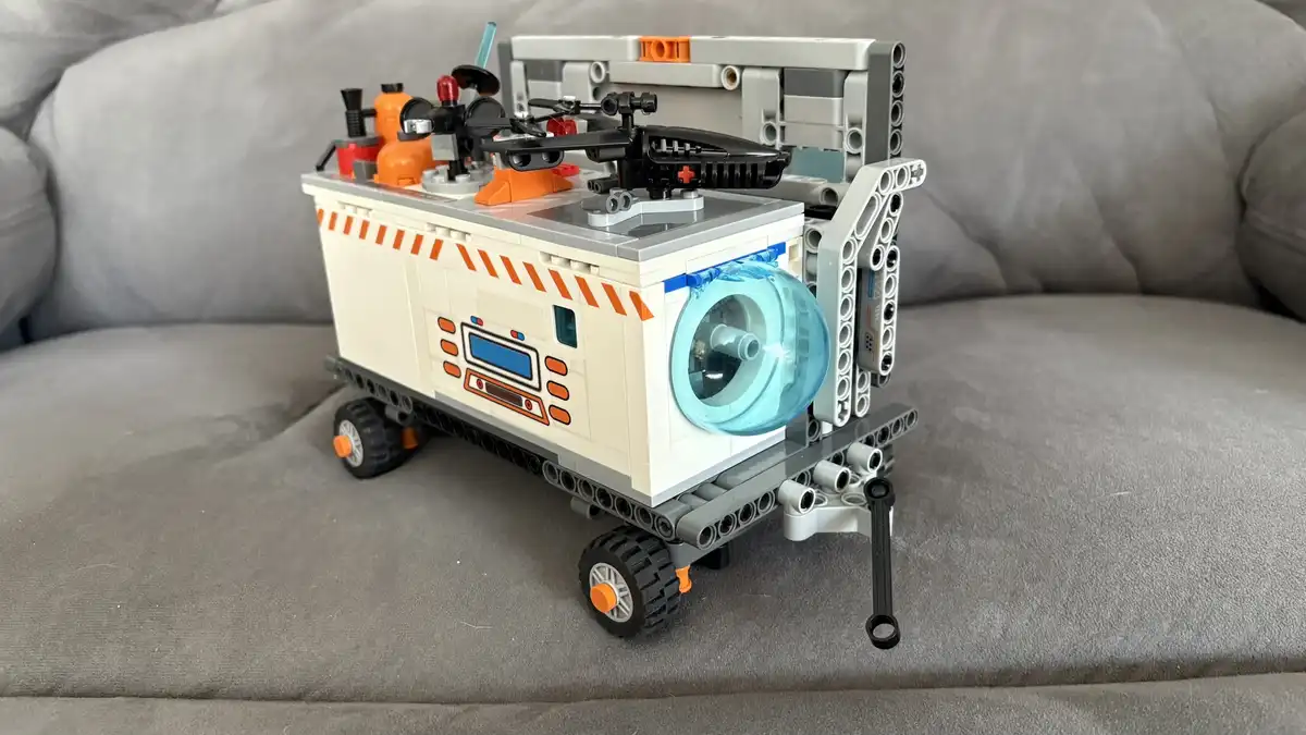A detailed LEGO model of a futuristic vehicle, resembling a mobile lab or medical transport, sits on a gray couch. It…