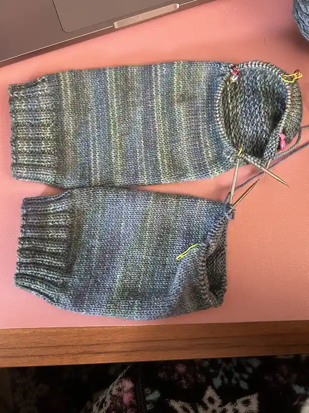 Two hand-knitted socks in progress, made from multicolored yarn with red and yellow stitch markers, lie on a pink surface…