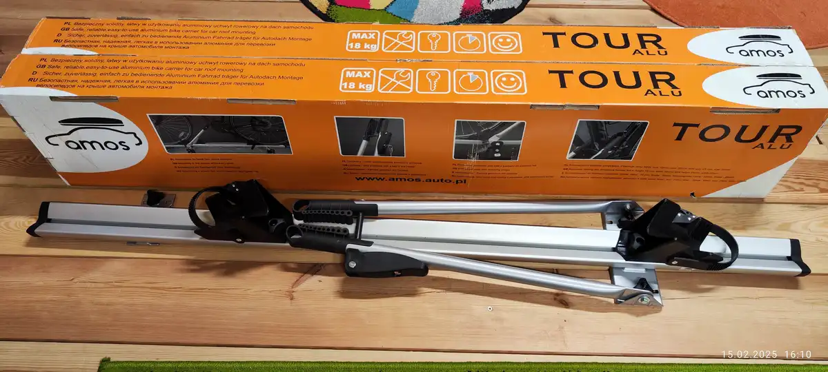 An unboxed silver and black aluminum bicycle roof rack, model 'TOUR ALU' by 'amos', lies on a wooden floor in front of its…