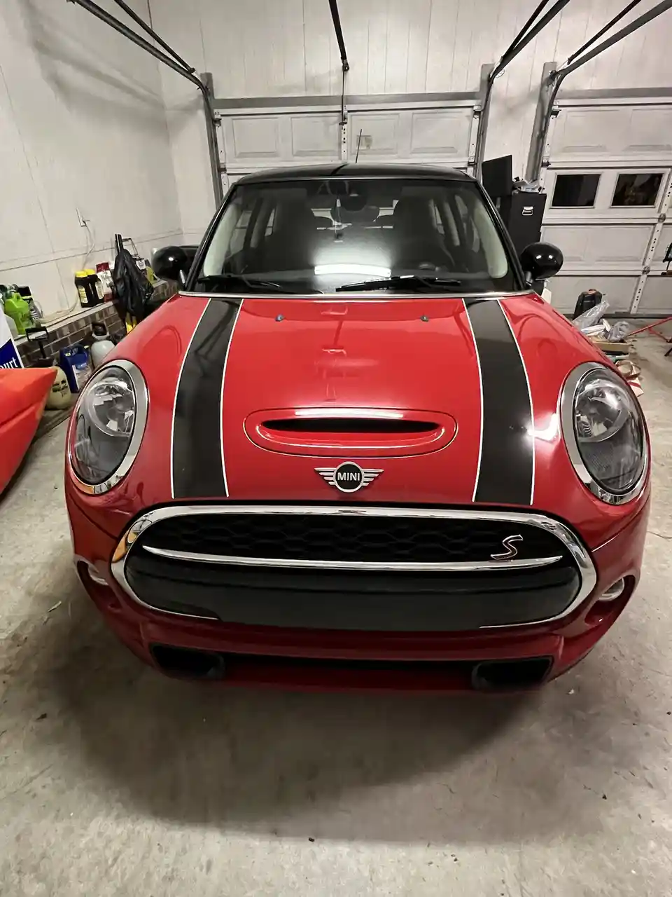 A red Mini Cooper S with black racing stripes is parked inside a cluttered garage, facing the camera. The car's hood…