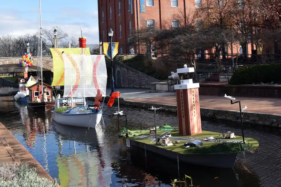 A canal features two decorated boats: one with baseball-themed sails and another floating island with a miniature airport…