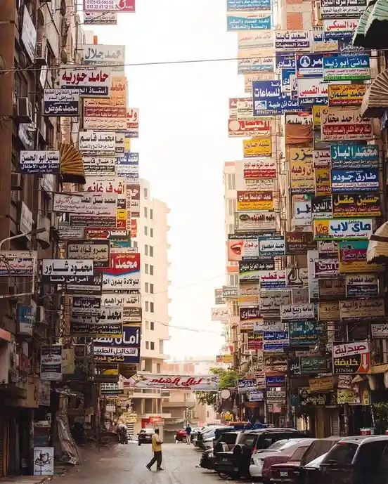 A narrow urban street is densely packed with colorful Arabic business signs hanging from buildings on both sides, creating a…