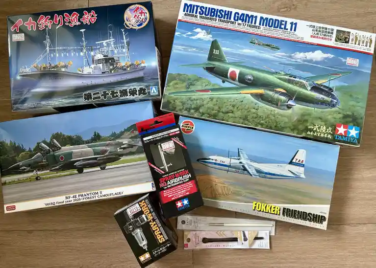 A flat lay of several model kits on a wooden surface, including a squid fishing boat, a Mitsubishi G4M1 aircraft, an RF-4E…