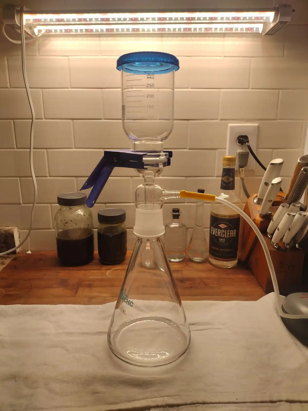 A makeshift distillation apparatus made from glassware, a blue-lidded measuring cup, and a clamp, sits on a kitchen counter…