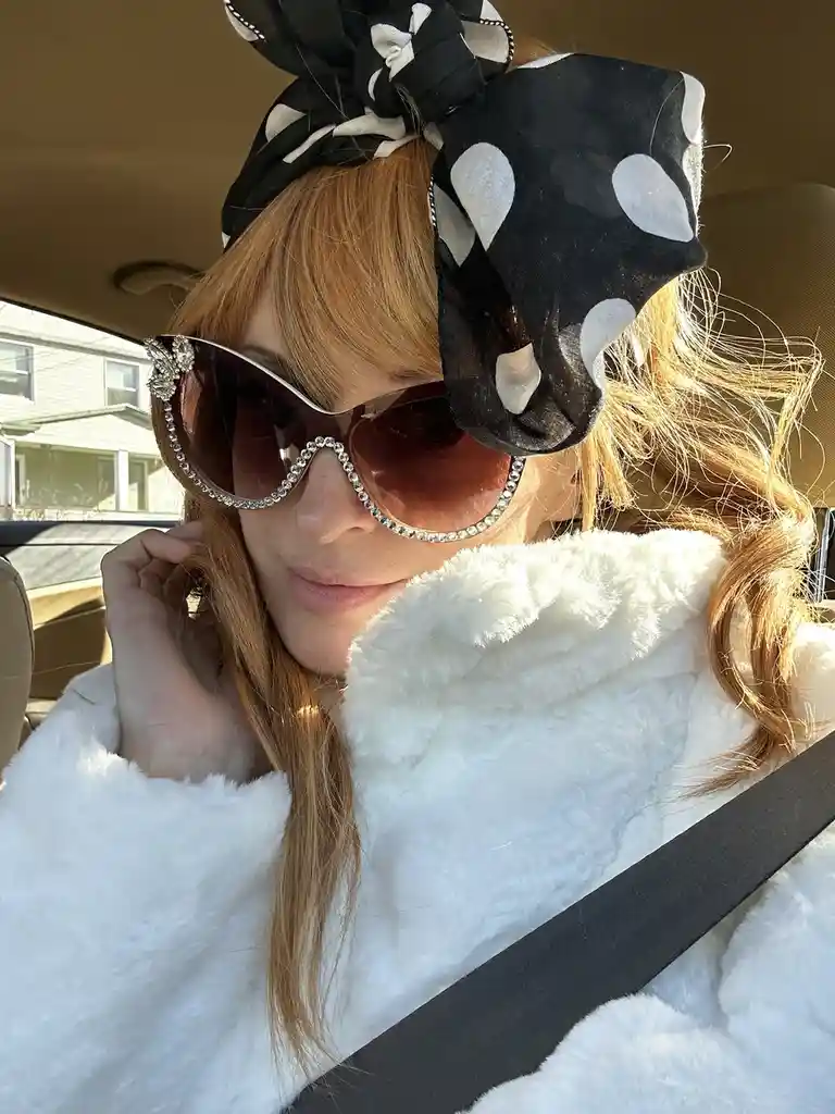 A person with reddish-blonde hair sits in a car wearing a large black-and-white polka dot headscarf, oversized sunglasses…