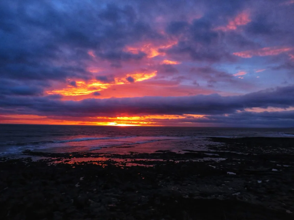A dramatic sunset over a rocky shoreline, with fiery orange and red clouds against a deep purple sky, reflecting on the…