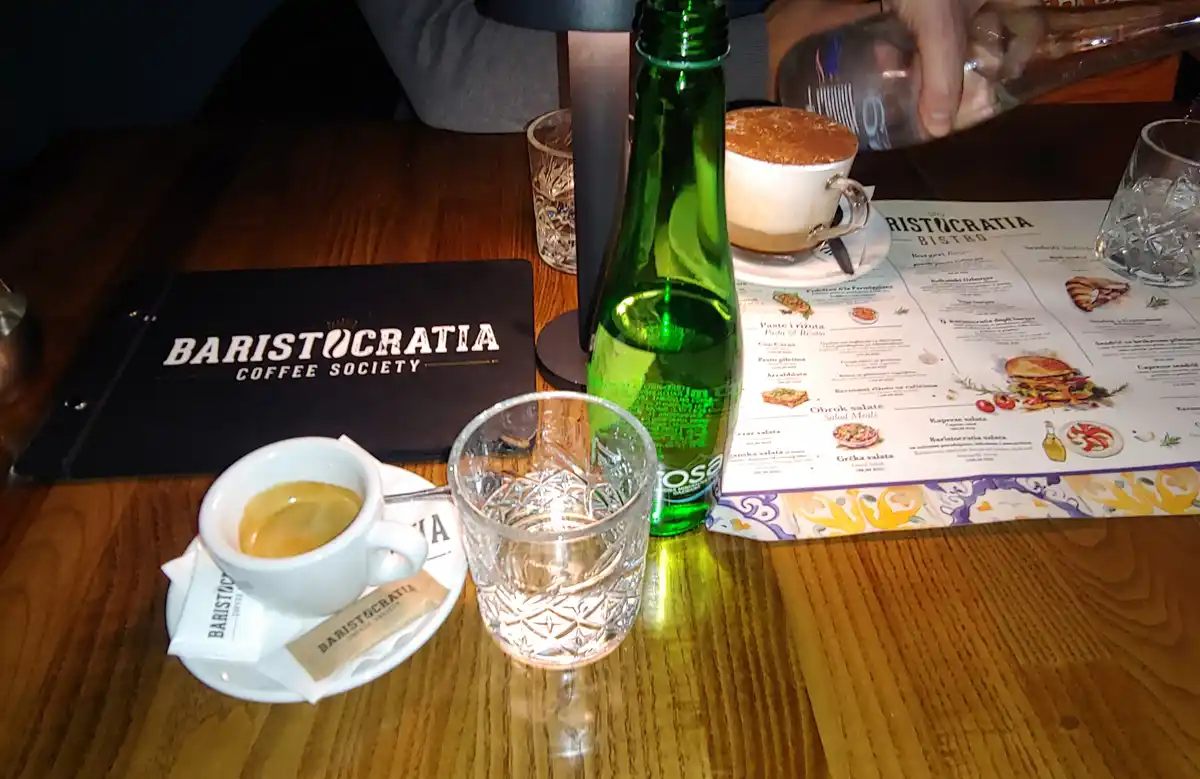 A wooden table at Baristocratia Coffee Society holds an espresso, a green bottle, a water glass, and a menu. A hand pours…