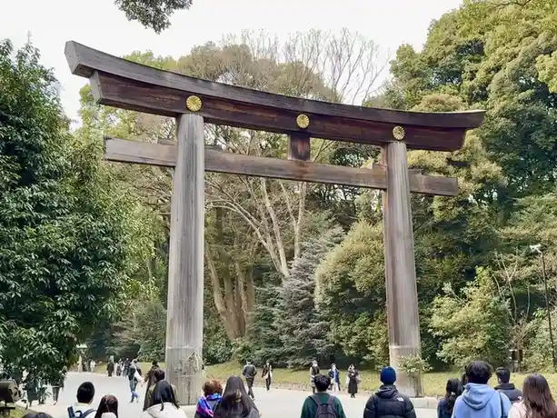 a large wooden torii gate with golden circular ornaments stands in a park surrounded by trees, with numerous people standing…