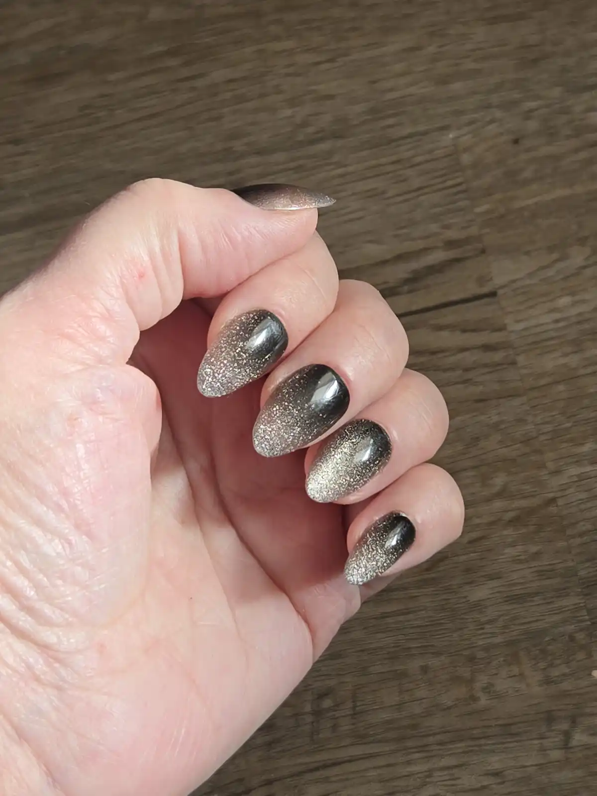 A close-up photo shows a person's hand with long, almond-shaped nails painted in a dark, glittery polish that transitions…