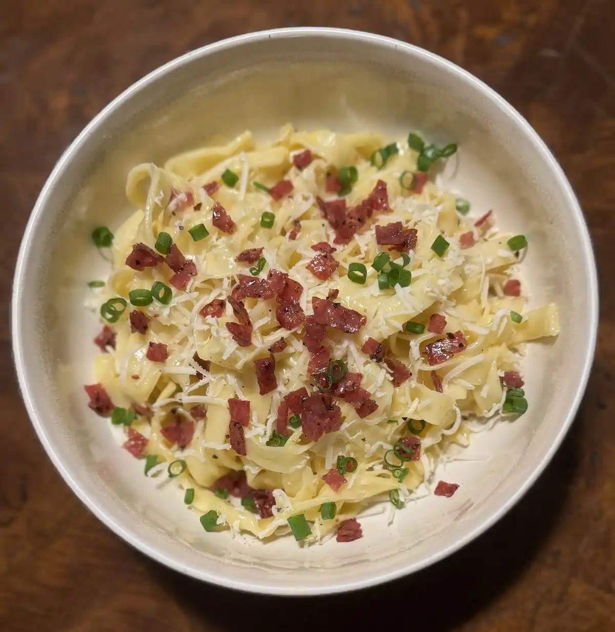 A top-down photo of a white bowl filled with wide pasta noodles coated in a creamy sauce, topped with shredded white cheese…