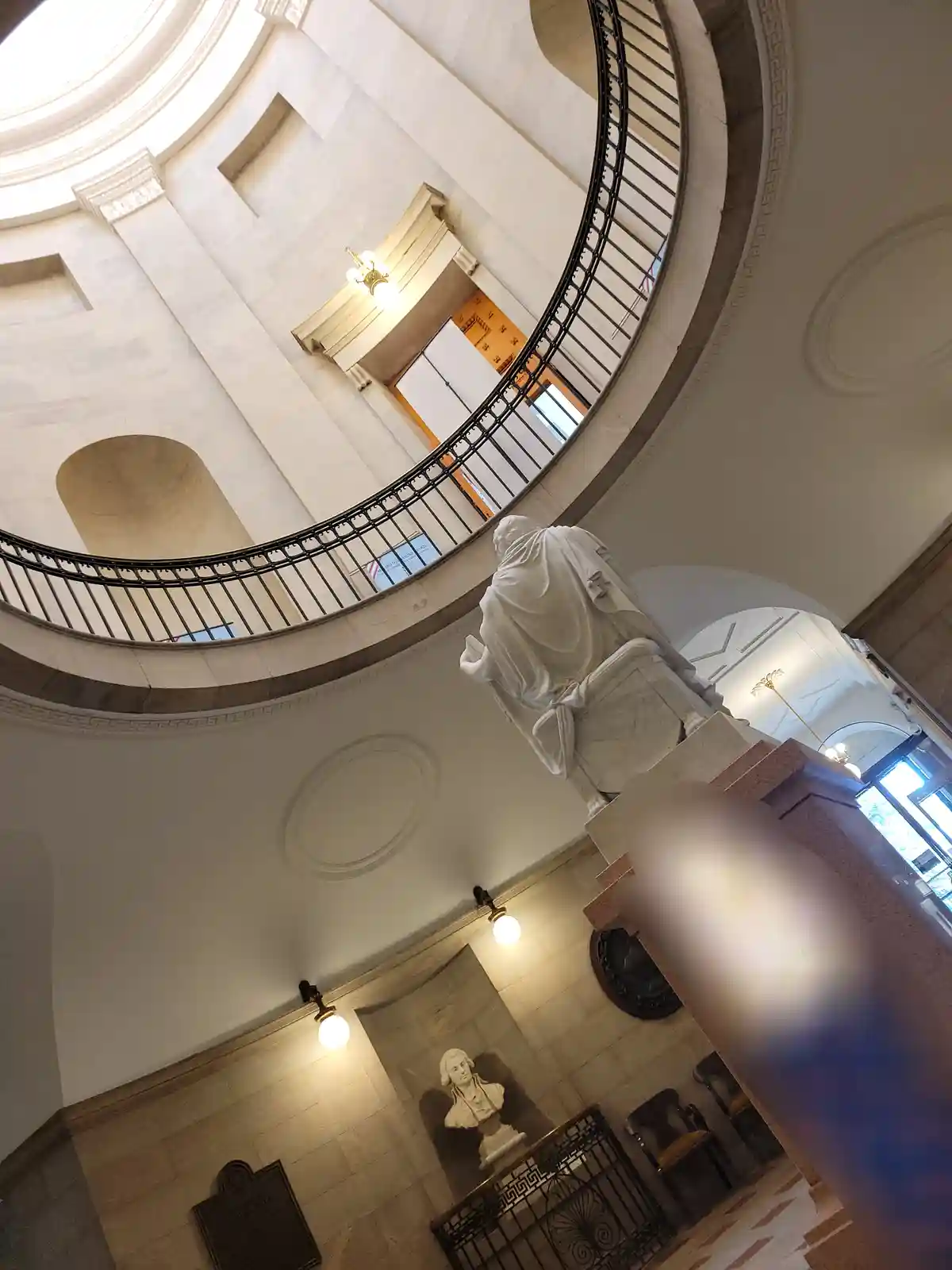 A low-angle photo looking up at a grand, circular interior with a marble statue of a robed figure on a pedestal. A black…