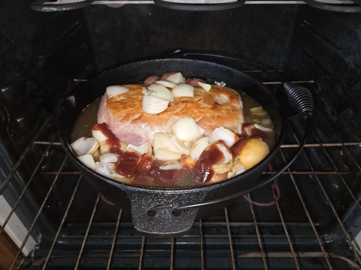 A close-up photo shows a large piece of ham roasting in a black cast-iron skillet inside an oven, surrounded by chopped…