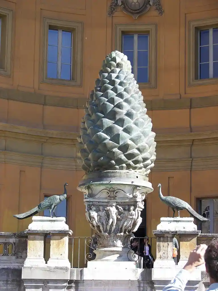 a large, ornate bronze statue shaped like a giant pinecone stands in front of a classical building with windows, flanked by…