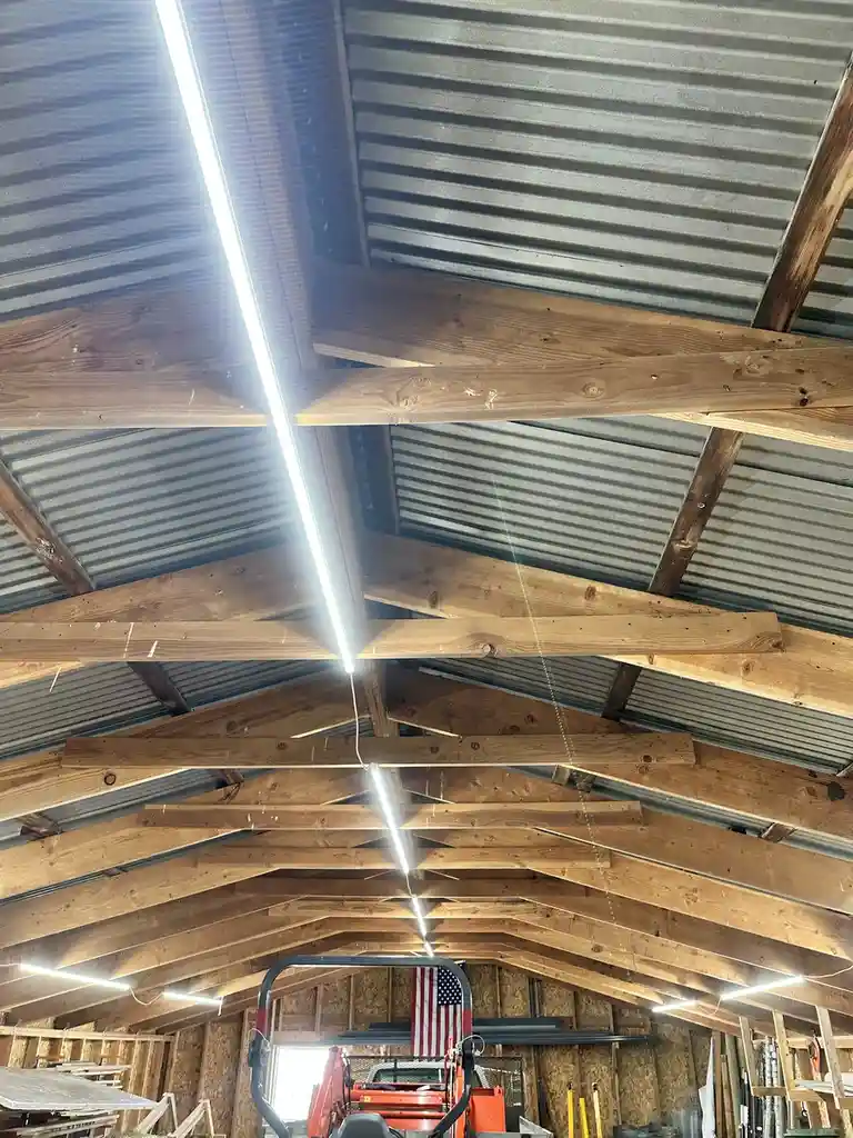 A low-angle photo looking up at the ceiling of a wooden-framed barn or workshop with corrugated metal roofing. Several long…