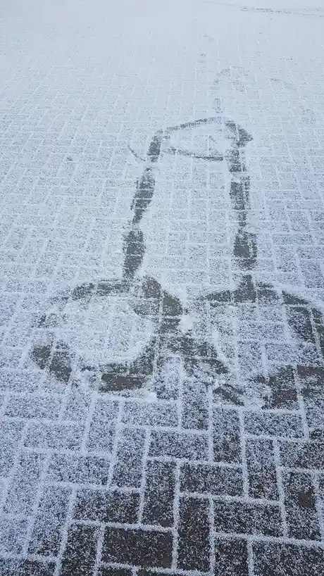 A snowy sidewalk shows a faint, dark outline of a person sitting on a chair, with the chair's legs and seat visible through…