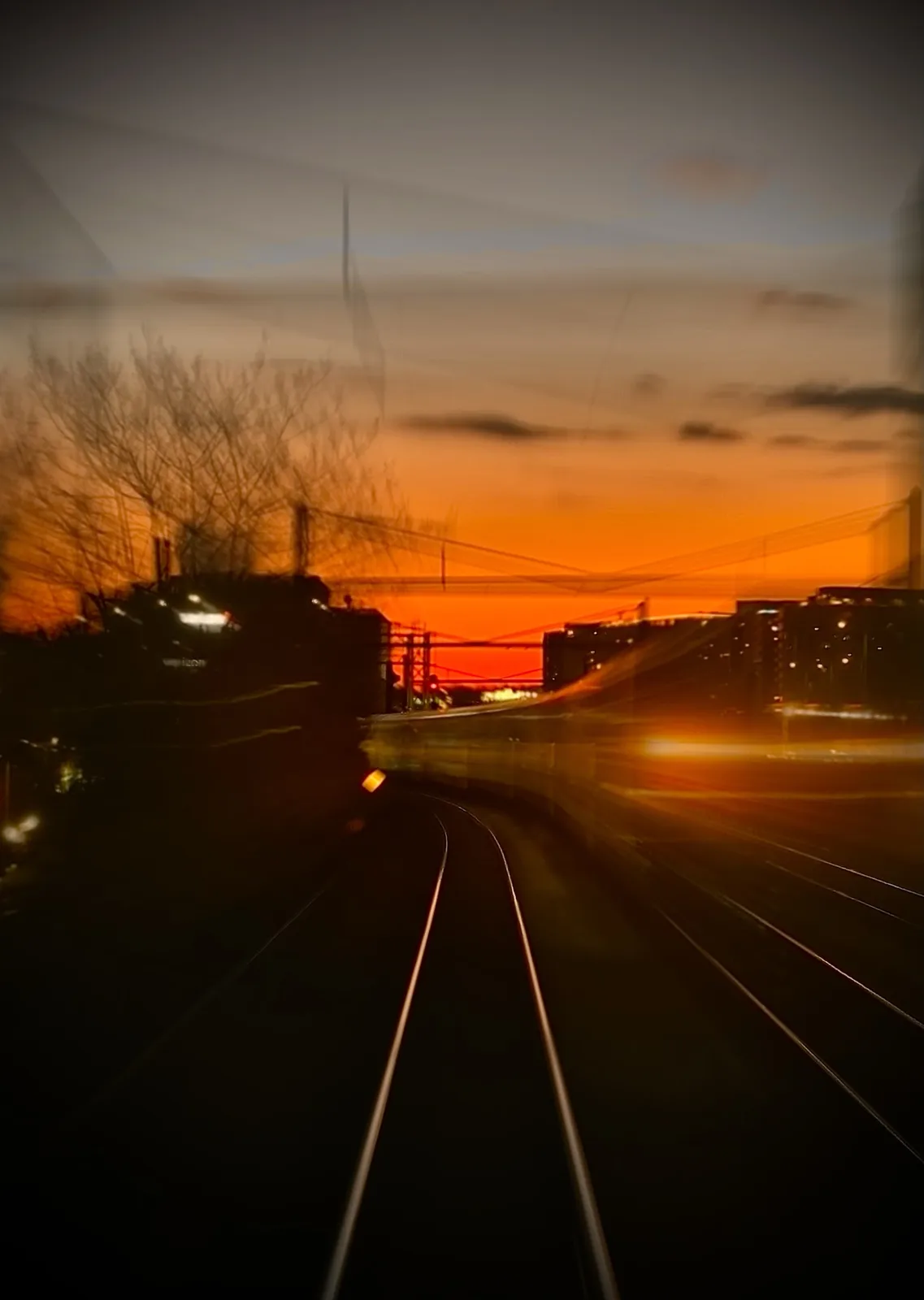 A motion-blurred photograph taken from a moving train at sunset, showing orange sky, distant buildings, and blurred tracks…
