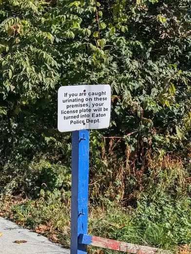 A white rectangular sign on a blue post stands beside a sidewalk, surrounded by green bushes and grass. The sign reads: 'If…