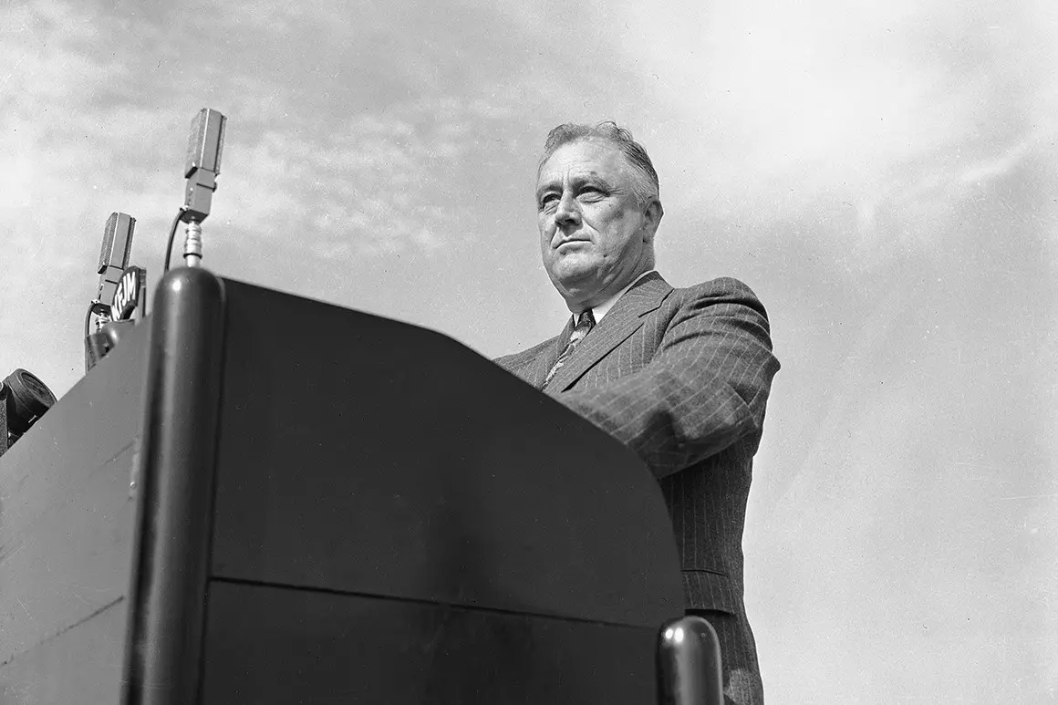 a black-and-white photo shows a man in a suit standing at a podium with microphones, looking off to the side with a serious…