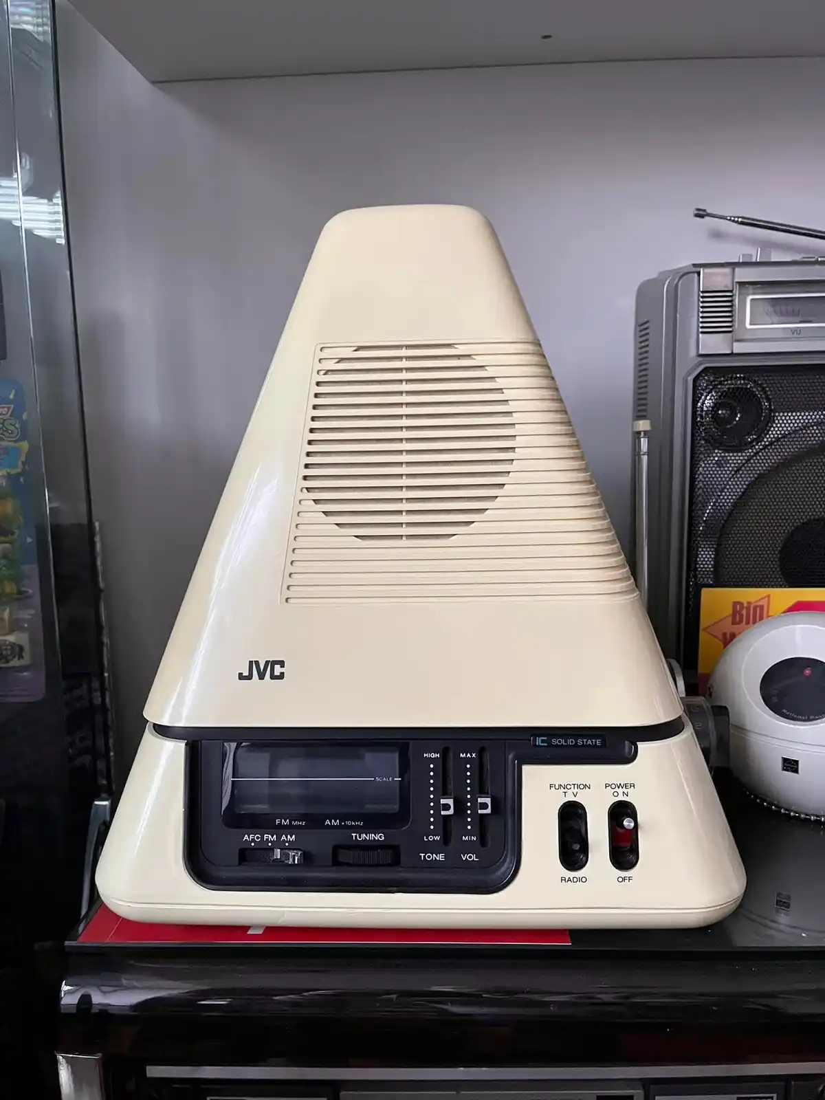 A cream-colored, pyramid-shaped JVC vintage radio with a black control panel featuring dials and switches for tuning, tone…