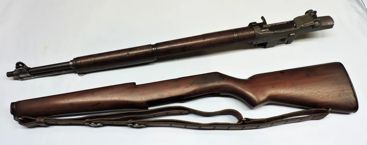A disassembled bolt-action rifle with wooden stock and metal barrel lies on a white surface, accompanied by its leather…