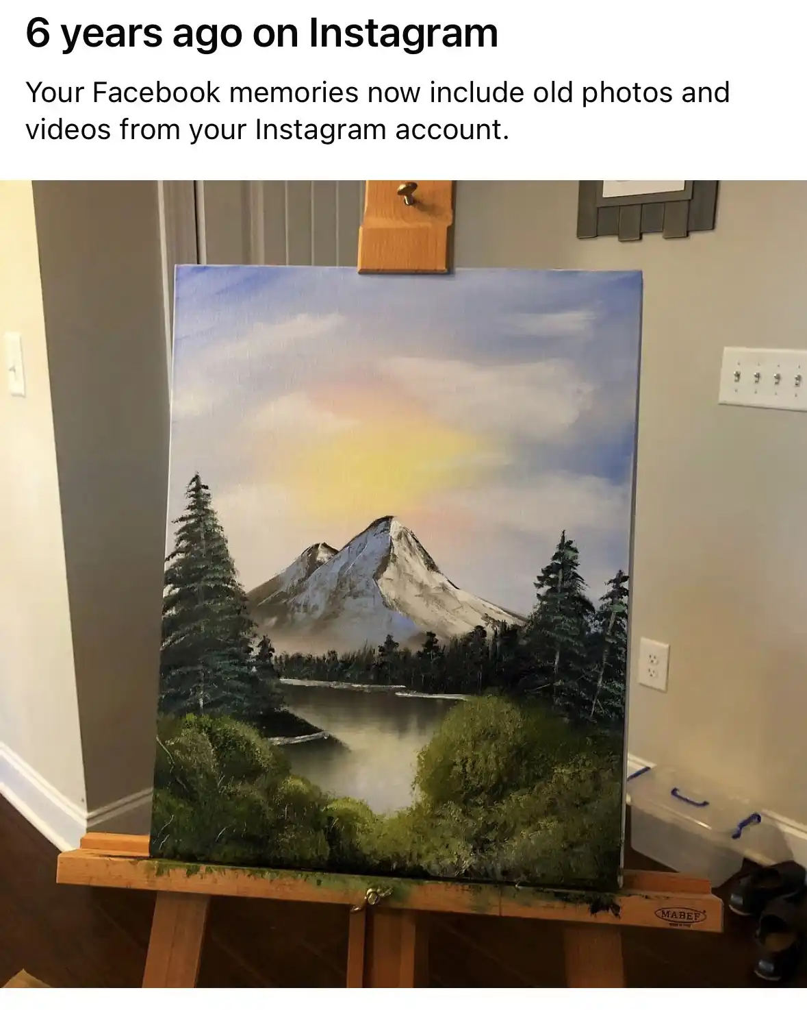 A painting of a snowy mountain landscape with evergreen trees and a lake is displayed on a wooden easel in a room. The…