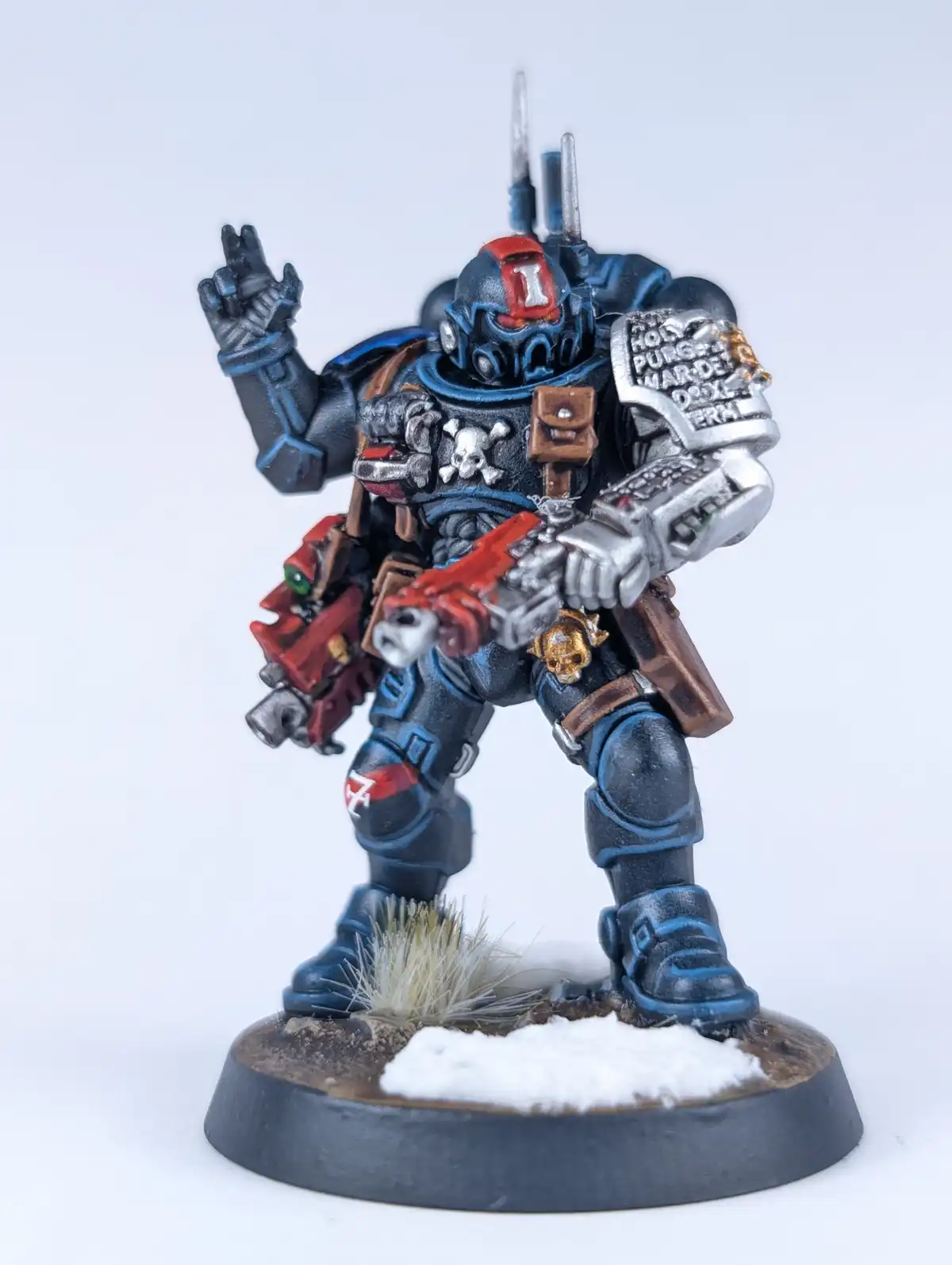 A painted miniature of a Space Marine in dark blue and red armor, holding a weapon and making a hand gesture, stands on a…