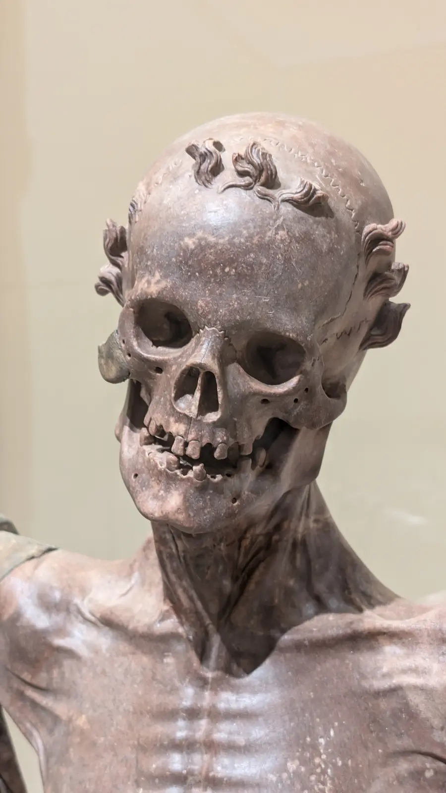 A close-up of a brownish, marble-like sculpture of a human skull and upper torso, with stylized hair carved on the head and…