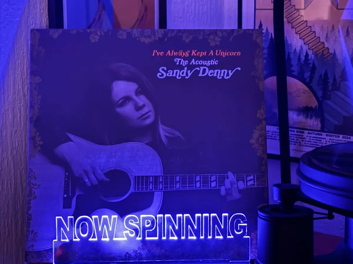 A vinyl record album cover for Sandy Denny's 'I've Always Kept A Unicorn: The Acoustic' is shown under blue light, with a…