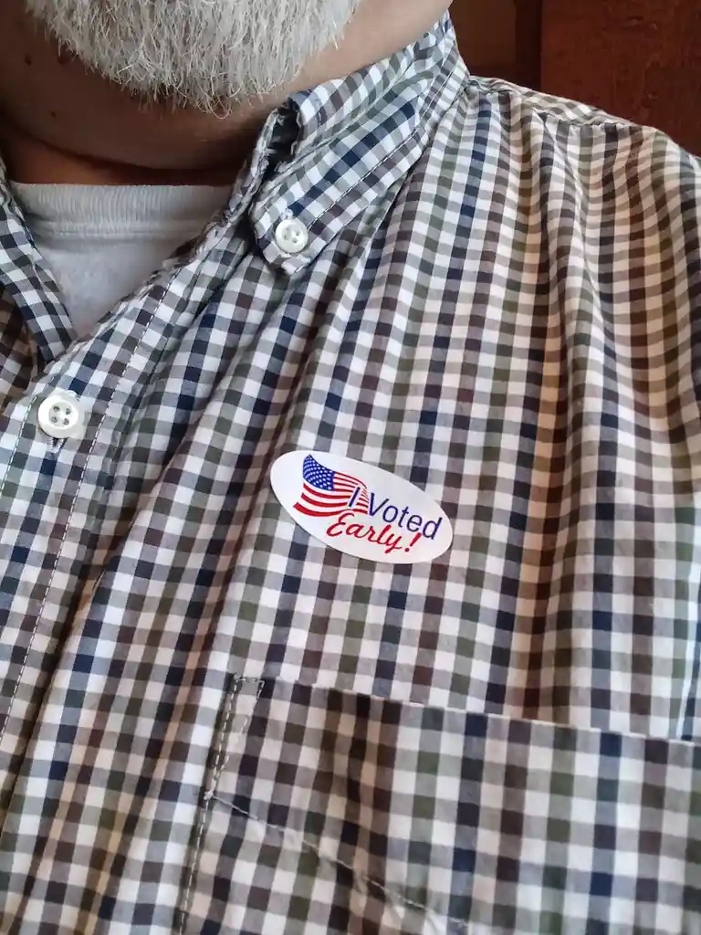 A close-up photo shows a person wearing a plaid button-up shirt with a white oval sticker pinned to the chest. The sticker…