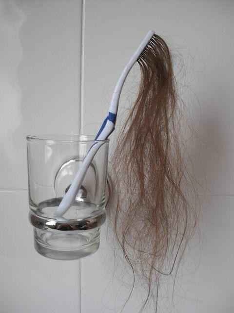 A white and blue toothbrush is stuck vertically in a clear glass cup mounted on a tiled wall, with a long, tangled clump of…