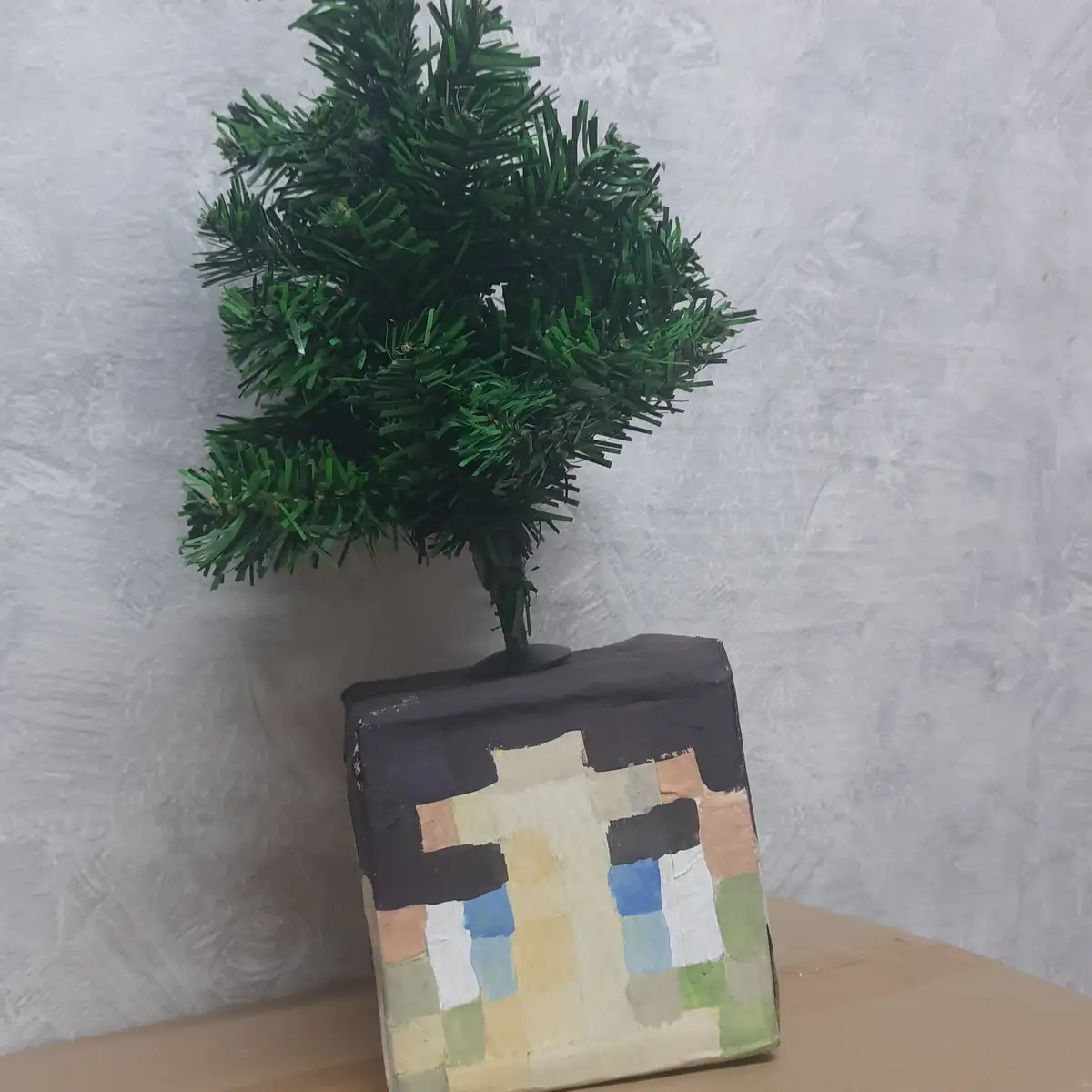 A small artificial green Christmas tree is planted in a handmade, blocky, pixelated vase that resembles a Minecraft…