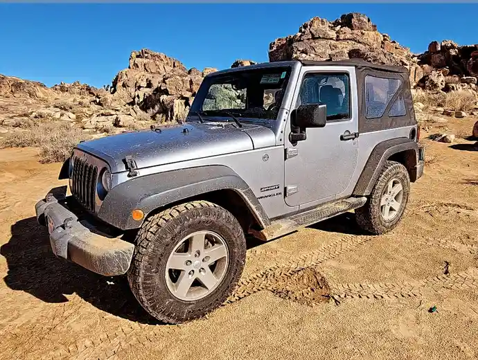 A silver Jeep Wrangler Sport is parked in a desert with rocky formations in the background, covered in dust and dirt. The…