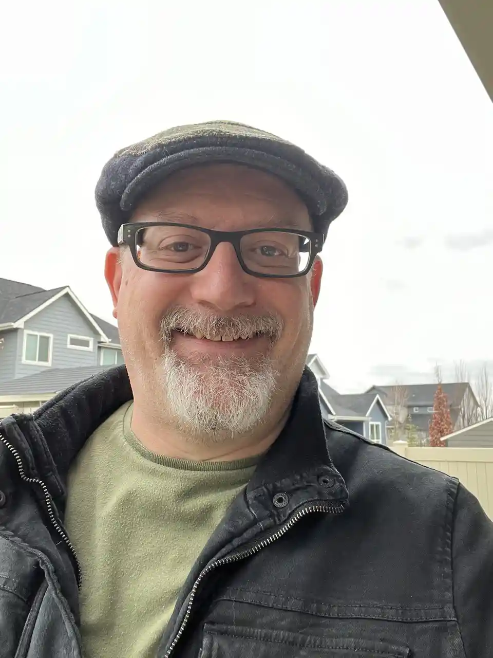 A middle-aged man with glasses and a flat cap smiles at the camera while wearing a black jacket over a green shirt, standing…