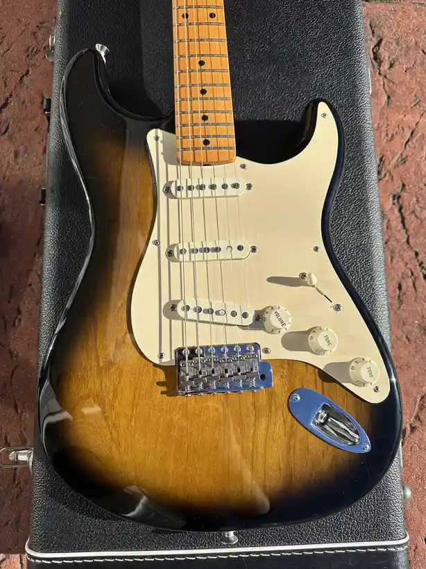 A sunburst-finish electric guitar with a maple neck and white pickguard rests inside a black case on a red brick surface…