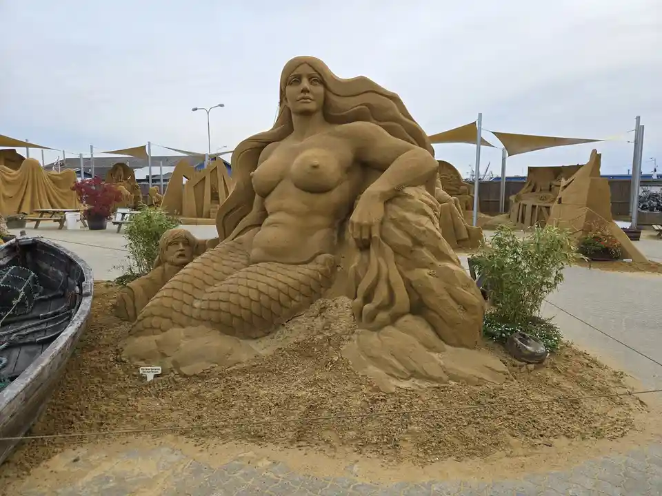 A large, detailed sand sculpture of a mermaid with long hair and a scaled tail sits prominently in an outdoor sand art…