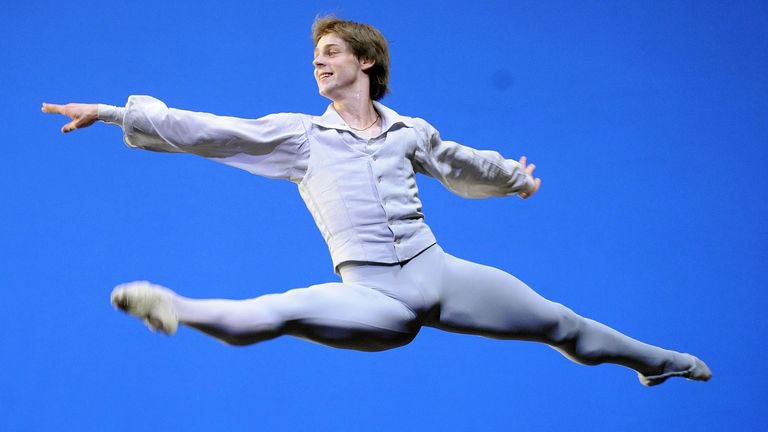 A male ballet dancer in a light gray costume is captured mid-air performing a split leap against a solid blue background. He…