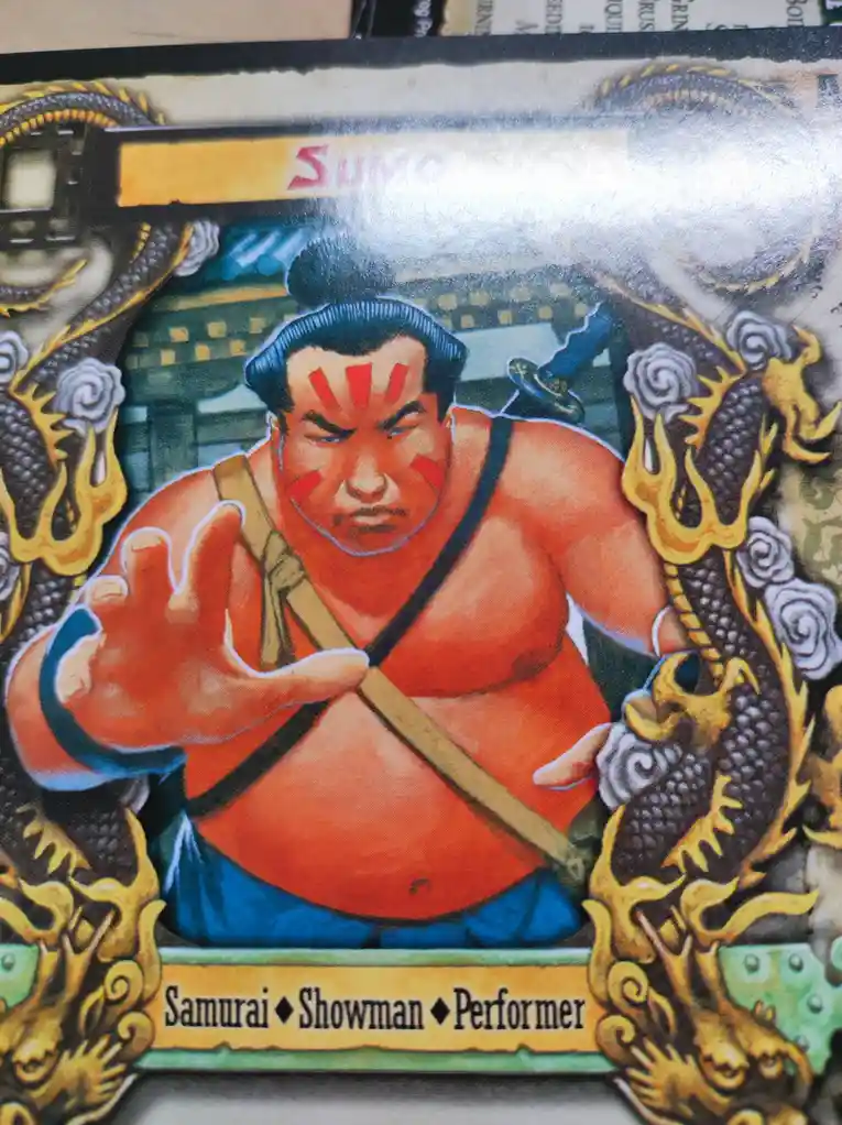 A colorful illustrated trading card depicts a muscular sumo wrestler with red face paint, wearing a mawashi, and surrounded…