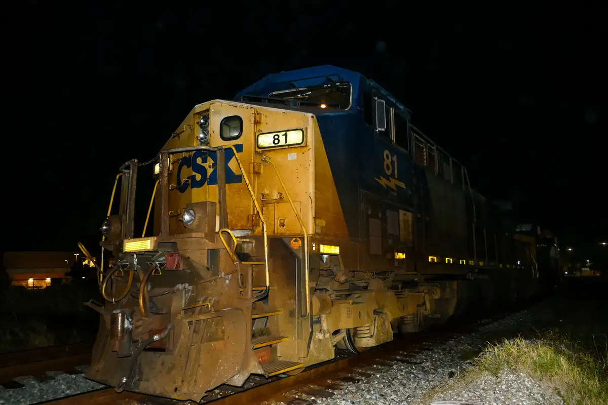 A nighttime photo of a CSX freight train locomotive, number 81, with blue and yellow livery, sitting on tracks. The number…