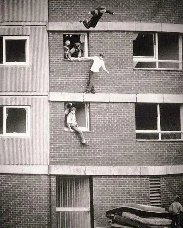 A black-and-white photo shows several children on a brick apartment building. One child is mid-air, having jumped from a…